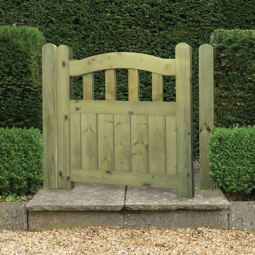 The KDM Omega Top Regency Euro-Gate, featuring vertical planks and an arched top, stands between trimmed green hedges on stone paving with gravel in the foreground.