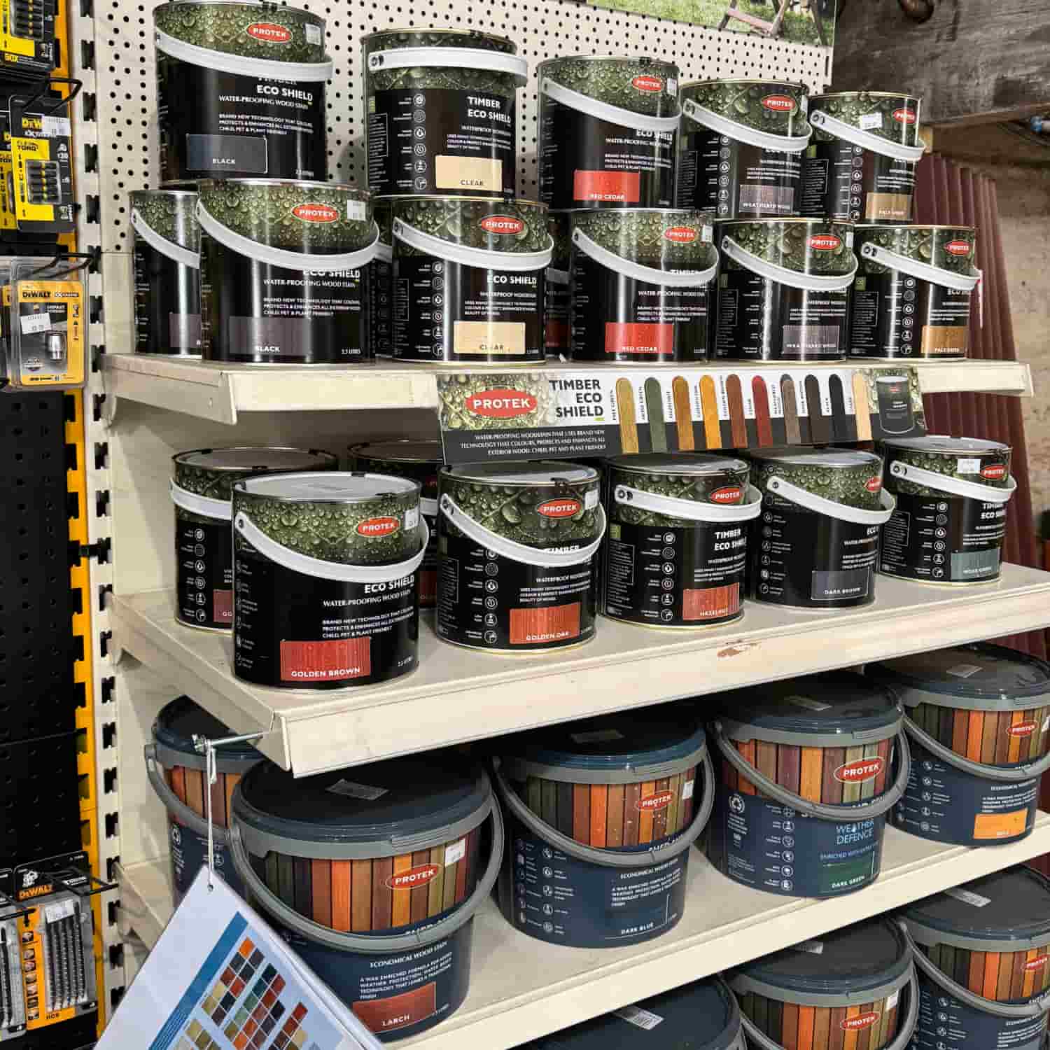 Shelves stocked with various cans of Protex wood stain and protective coatings in different finishes and colors, displayed in a hardware store. Color samples hang on the side and front of the shelves.