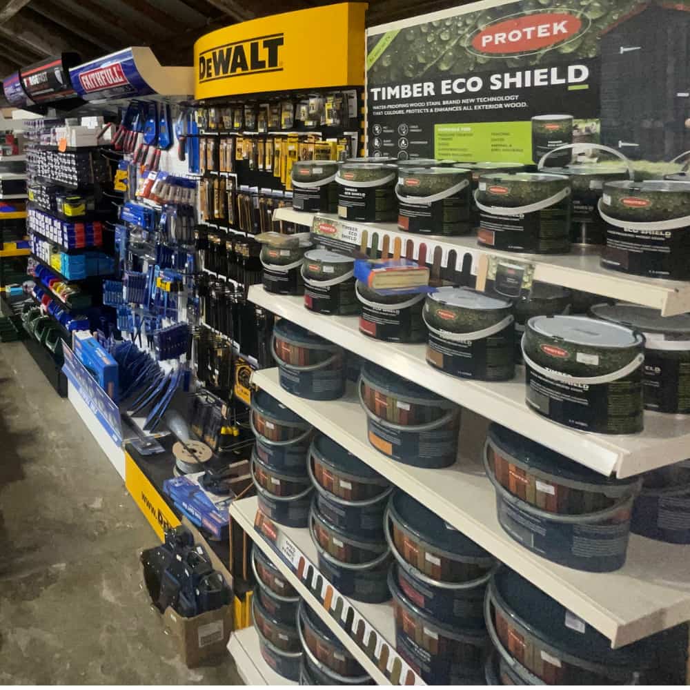 Shelves in a hardware store display various tools, paintbrushes, and tubs of Protek Timber Eco Shield wood treatment. DeWalt tools are also visible, and the aisles are neatly organized with products.