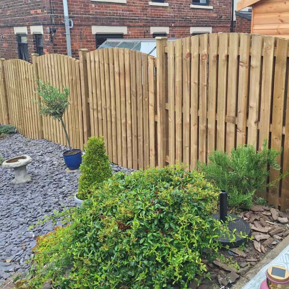 A wooden arched fence separates two gardens; in the foreground, there are various green bushes and shrubs planted in a gravel bed with slate stones. A small birdbath is also visible.