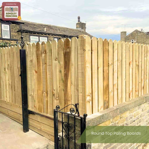 A tall fence of Bingley Fencing Round Top Paling Boards PSE (75mm x 19mm) stands by a low stone wall and black metal gate, with houses behind—an elegant example of quality picket fencing.