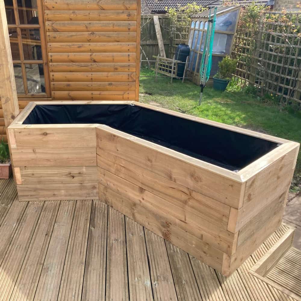 A large, L-shaped wooden planter with a black liner inside is placed on a wooden deck in a garden, near a wooden shed and surrounded by fencing and plants.