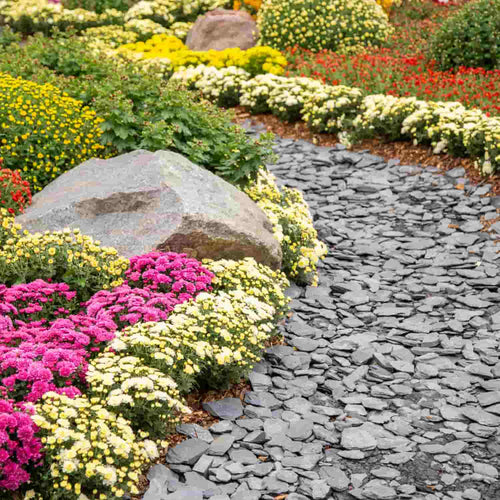 A curving stone path winds through a vibrant garden with green plants, large rocks, and The Heritage Stone Company’s Plum Slate - 40mm Pieces - 20kg, bordered by blooming pink, yellow, white, and red flowers.