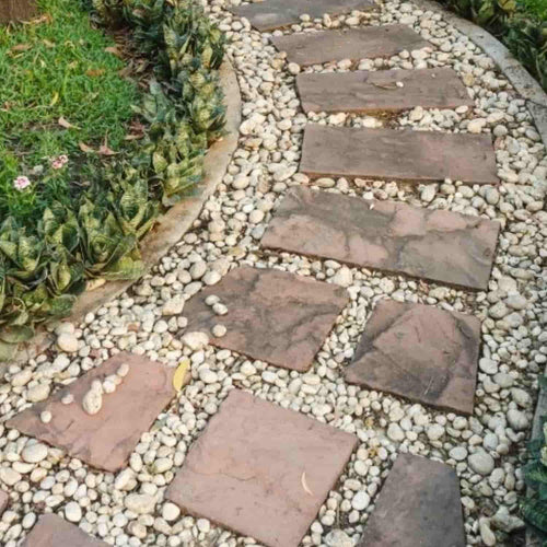 A curved garden pathway composed of large, rectangular stone slabs is elegantly set in a bed of 40mm pieces from The Heritage Stone Company's Blue Slate - 20kg. On the left side, a border of green plants with jagged leaves runs alongside a grassy area, enhancing the decorative garden ambiance.