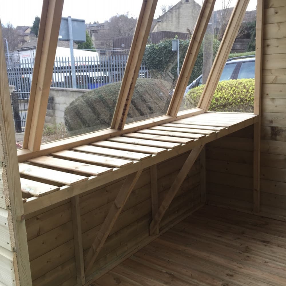 A wooden greenhouse interior with large slanted windows, a sturdy wooden bench beneath the windows, and wooden floorboards. Bushes and a metal fence are visible outside.