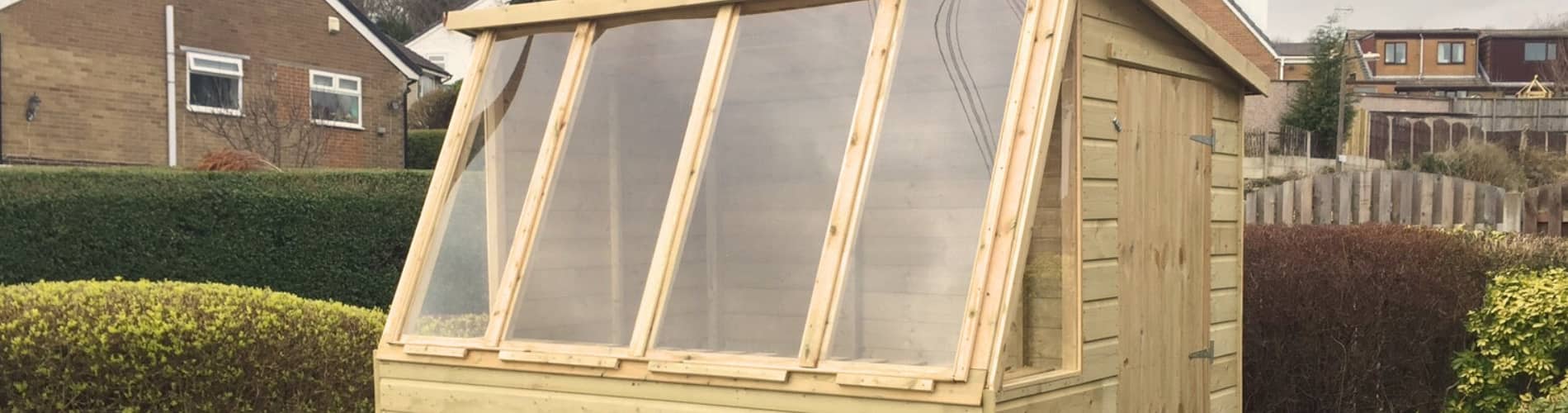 A small wooden greenhouse shed with a sloped roof and large clear plastic windows, situated in a garden with hedges and neighboring houses in the background.