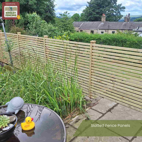 A garden with tall grass features a round table with a plant and yellow rubber duck, and a contemporary KDM Slatted Fence Panel in a light color. Trees and houses can be seen beyond. Text reads: Slatted Fence Panels.