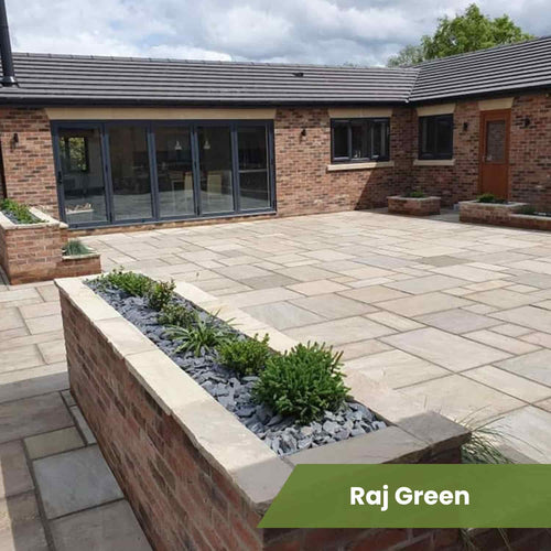 A modern patio features Bingley Fencing’s Indian Sandstone Paving Project Pack with raised brick planters filled with small green plants and grey stones. A brick house with large glass doors is in the background. Text overlay reads Raj Green.