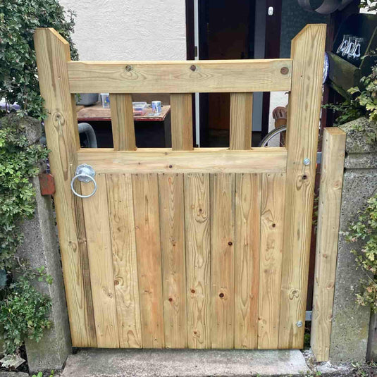 The KDM Regency Euro-Gate is a timber garden gate with vertical planks, three rectangular openings at the top, and a metal ring latch. This classic gate attaches to stone posts and is partially surrounded by greenery.