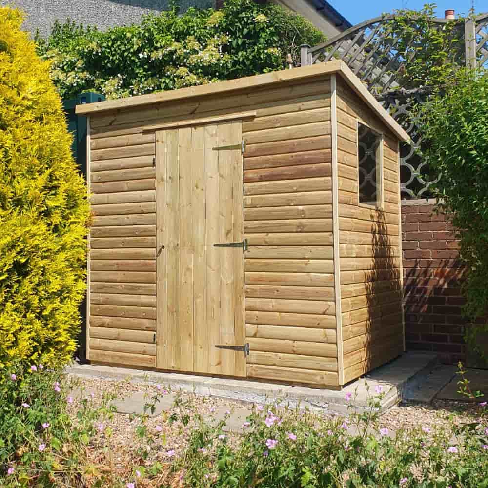 A small wooden garden shed with a single door and window sits on a paved area, surrounded by green shrubs, flowers, and a brick wall in the background.