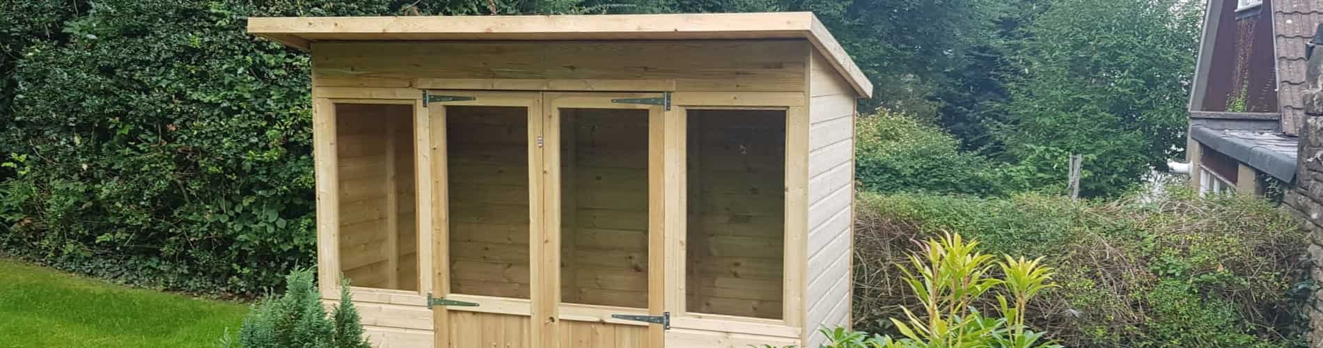 A wooden garden shed with a slanted roof, three glass-paneled doors, and surrounded by greenery, bushes, and plants, positioned near a hedge and a house exterior.