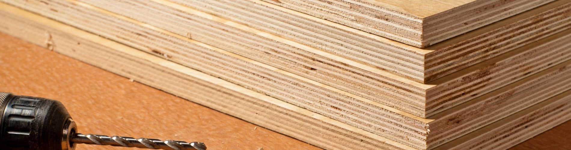 A stack of neatly cut plywood sheets sits on a work surface next to a cordless drill with a drill bit attached, showcasing woodworking materials and tools.