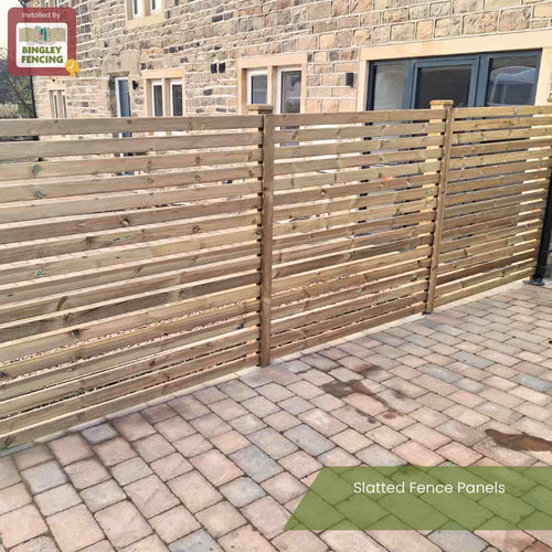 A KDM Slatted Fence Panel with a modern slatted design stands on a paved patio beside a stone building, offering partial privacy. It features a Slatted Fence Panels label and KDM branding in the top left corner.