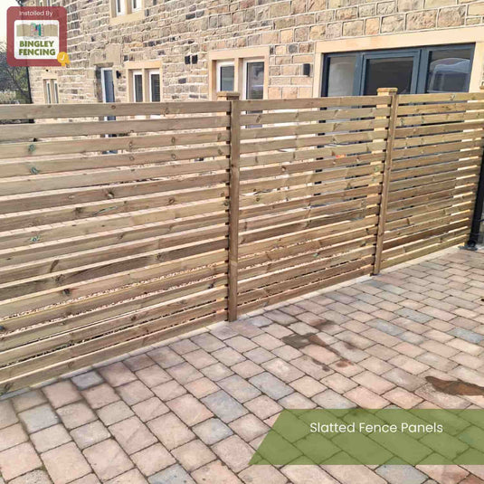 A KDM Slatted Fence Panel with a modern slatted design stands on a paved patio beside a stone building, offering partial privacy. It features a Slatted Fence Panels label and KDM branding in the top left corner.