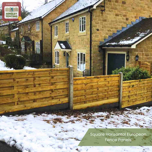 A house with tan brick and dark trim stands behind a wooden fence of KDM Square Horizontal Fence Panels (SH). The solid horizontal slats enclose a snowy yard, while snow covers the ground and parts of the road.