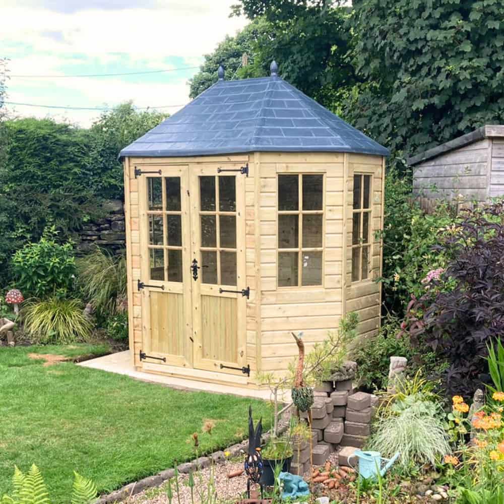 A small, light wooden garden gazebo with glass windows and a blue roof stands on a stone base, surrounded by green plants and a lawn in a lush garden.