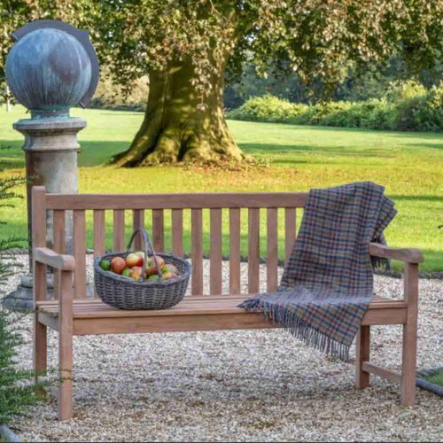 The 1.5m Teak Garden Bench by Bingley Fencing sits on gravel, adorned with a plaid blanket on one arm and a basket of fruit on the seat, with green grass and a large tree in the background.