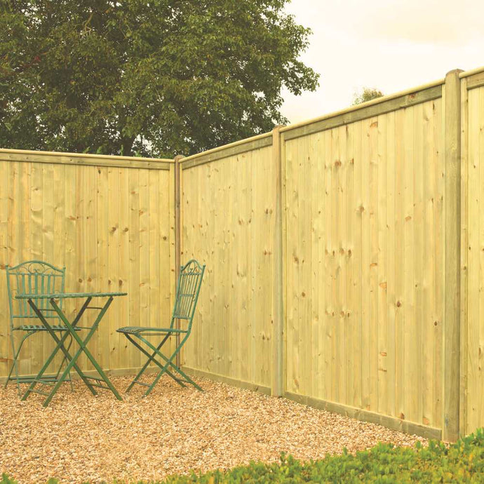 A small outdoor area features KDM Tongue & Groove European Fence Panels Flat Top for privacy, a gravel ground, and a green metal table with two matching chairs. Lush greenery and a tree can be seen beyond the fence.