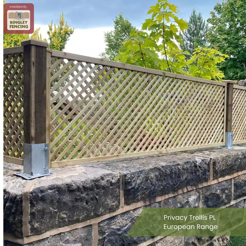 A wooden privacy trellis fence with a lattice design is mounted on a stone wall, surrounded by greenery and trees. A sign reads “Installed by Bingley Fencing.” Text at the bottom says “Privacy Trellis PL European Range.”.
