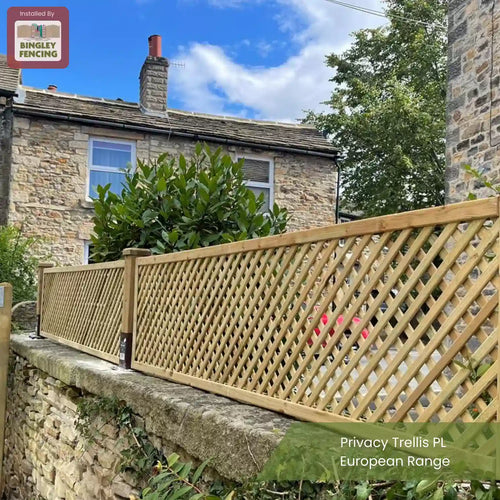A wooden lattice privacy trellis fence is installed on a stone wall outside a stone house with large windows and greenery. The sky is blue with some clouds. Text reads: “Privacy Trellis PL European Range.”.