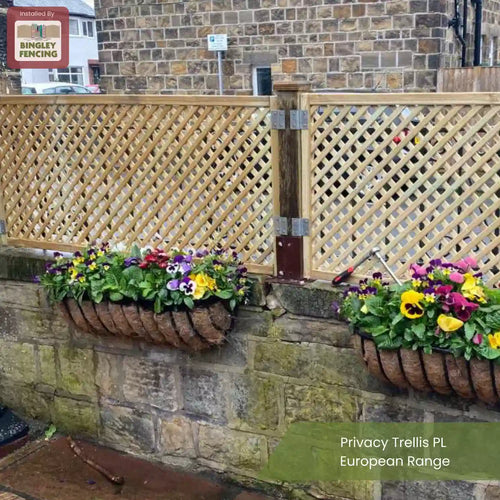 A wooden privacy trellis fence with lattice panels is installed atop a stone wall. Two colorful flower baskets hang on the wall below the fence. The setting is an outdoor garden area.