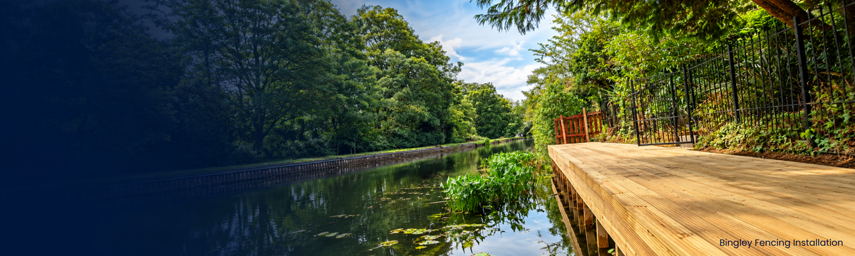 A wooden deck with metal fencing overlooks a calm canal lined with lush green trees under a partly cloudy sky.