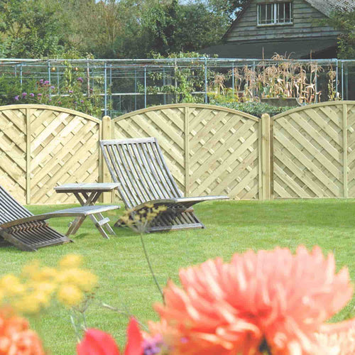 KDM 'V' Arched Solid Fence Panel - VA forms the backdrop for wooden garden chairs and a small table on a green lawn, with blooming flowers in front and a garden shed visible behind.