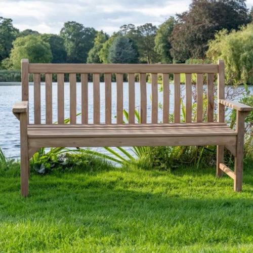 A Bingley Fencing 1.5m Wave Back Teak Garden Bench rests on lush green grass beside a serene lake, framed by trees and shrubs under a cloudy sky.