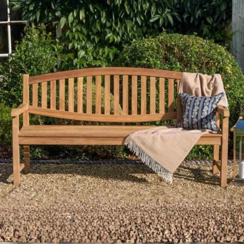 A 1.8m Curved Back Teak Garden Bench by Bingley Fencing sits on gravel with a beige fringed throw and a patterned pillow, set in front of green bushes.
