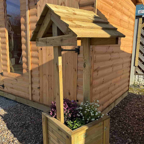 This Wishing Well Planter, crafted from pressure-treated timber and filled with purple and white flowers, sits on gravel beside a wooden cabin with log-style siding. Made by the staff at bingley fencing