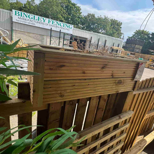 A Fence Top Planter – Wooden Decking & Balustrade Planter from Bingley Fencing, filled with dried plants, is mounted on a fence—serving as a practical and stylish fence planter ideal for any outdoor space. In the background, a sign displays “Bingley Fencing Manufacturers of Garden Fencing,” along with views of trees and fencing materials.