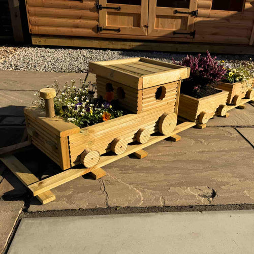 The Hand Crafted Wooden Train Planter by KDM, filled with vibrant flowers, sits on a stone patio near gravel and a wooden shed. Sunlight highlights this charming bespoke garden display.