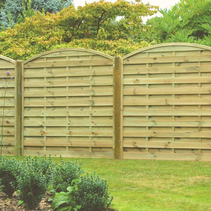 A KDM Arched Horizontal Fence Panel - AH180, made from pressure-treated timber, stands behind a green lawn and neat shrubbery, with tall leafy trees in the background.