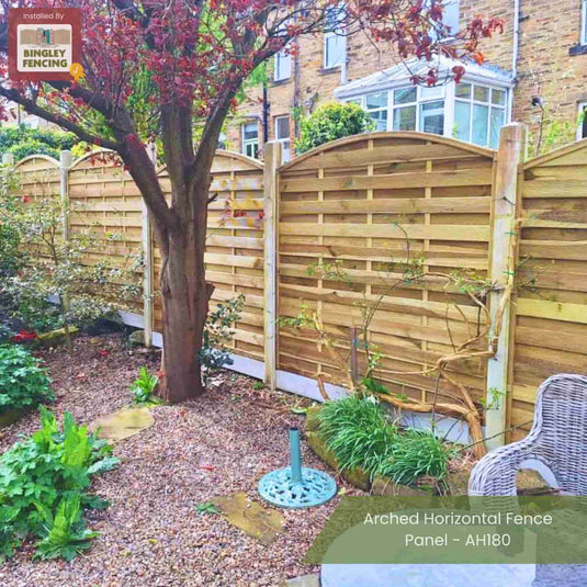 A garden with gravel paths, lush plants, a tree, a wicker chair, and tall KDM Arched Horizontal Fence Panels - AH180. Made from pressure-treated timber for elegance and durability.