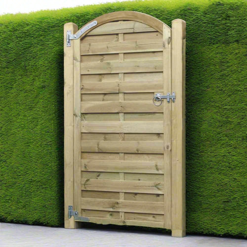 The KDM Arched Horizontal Euro-Gate (AHG180), featuring a curved top and metal hinges, is nestled in a dense, neatly trimmed green hedge. The paved ground enhances its charm, evoking the tidy, rustic entrance of a classic European garden.