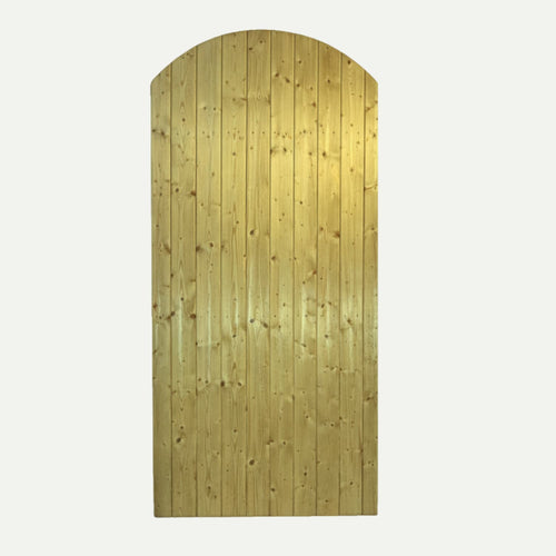 A tall, arched KDM Tongue & Groove Euro-Gate (TG180) stands closed, surrounded by a lush green hedge. Sunlight highlights the natural wood grain, creating a serene, garden-like atmosphere with gentle shadows.