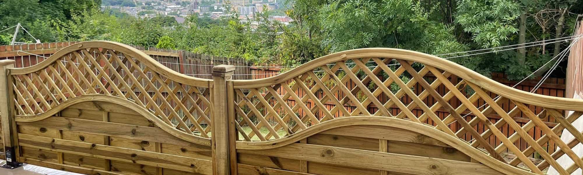Decorative wooden garden fence with a curved lattice top, set against greenery and overlooking a distant town or cityscape.