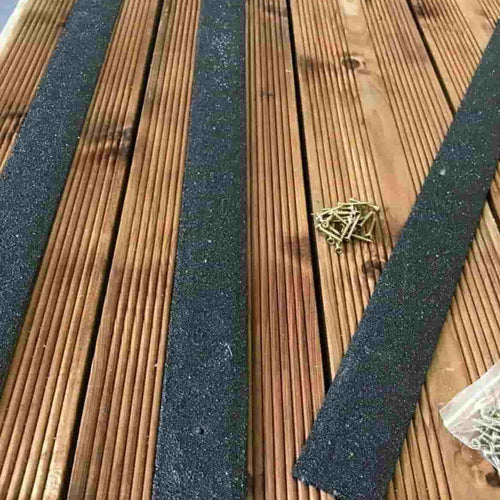 Three Bingley Fencing Anti Slip Decking Strips 1200mm Long–Black are placed on a wooden deck with gaps between planks, improving garden safety. Visible screws and a small plastic bag indicate installation in progress.