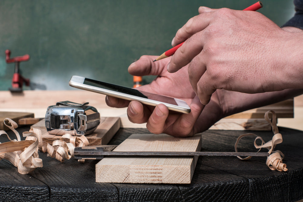 A person holding a smartphone in one hand and a pencil in the other, surrounded by woodworking tools and wood shavings on a workbench.