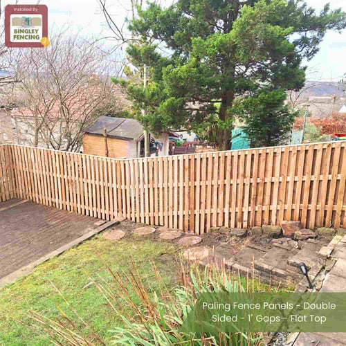 A sloped backyard with Bingley Fencing’s Paling Fence Panels—Double Sided, 1 Gaps, Flat Top—featuring evenly spaced vertical boards, set on a stone border near a wooden shed, trees, and decking.