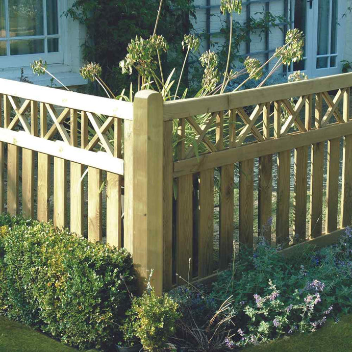The KDM Elite Cross Top Border Deck Panel (ECT90), made from tanalised softwood with a lattice design, stands surrounded by lush plants and flowers. The decorative top adds charm near a windowed building.