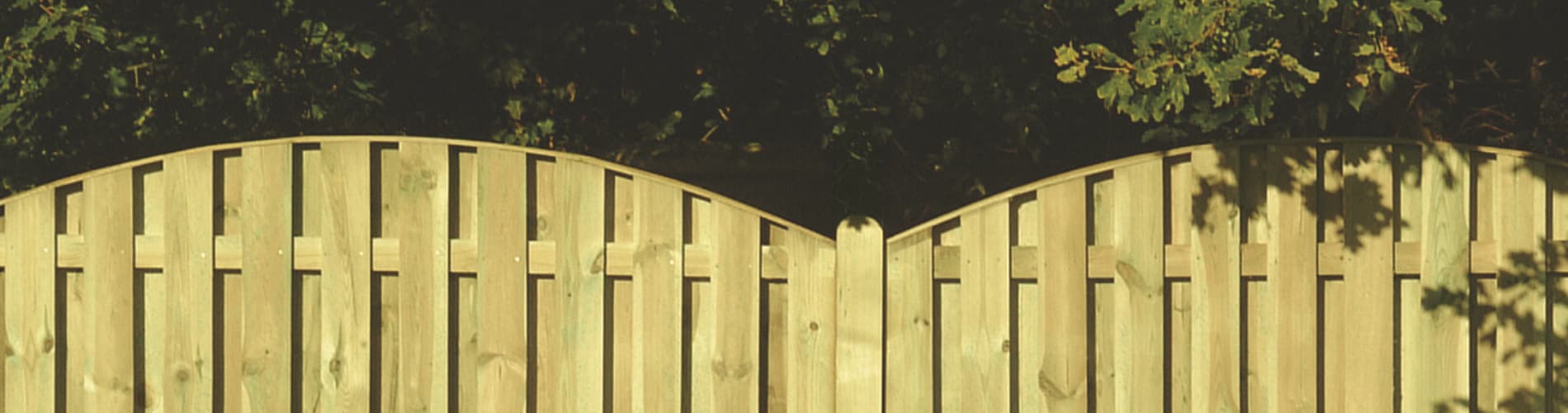 A light wooden fence with a gently curved top stands in front of leafy green trees, casting soft shadows on the fence in the sunlight.