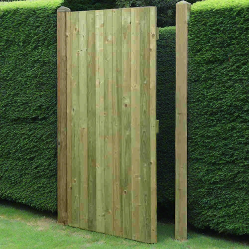 A KDM Tongue & Groove (Flat) Euro-Gate crafted from pressure-treated wooden planks stands amid a dense, manicured green hedge. It's surrounded by neatly trimmed grass and framed by two sturdy wooden posts, exuding a timeless charm.