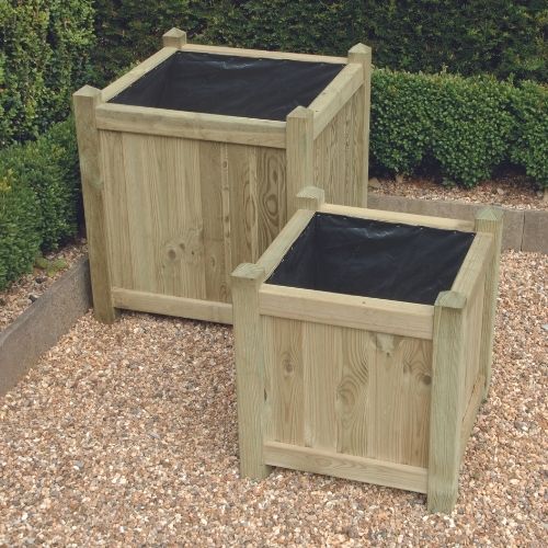 Two European Square Planters by KDM, available in custom sizes, are placed on a gravel surface. They feature a natural wood finish with black liners inside. Neatly trimmed green hedges surround the area, creating a garden-like atmosphere. Each planter arrives fully assembled for your convenience.