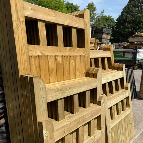 In a hardware store's yard, stacks of wooden Regency Euro-Gates by KDM are displayed outside. These light-colored wooden gates, featuring rectangular gaps, exude a Regency Style charm. Trees and stone buildings can be seen in the background under a sunny sky.