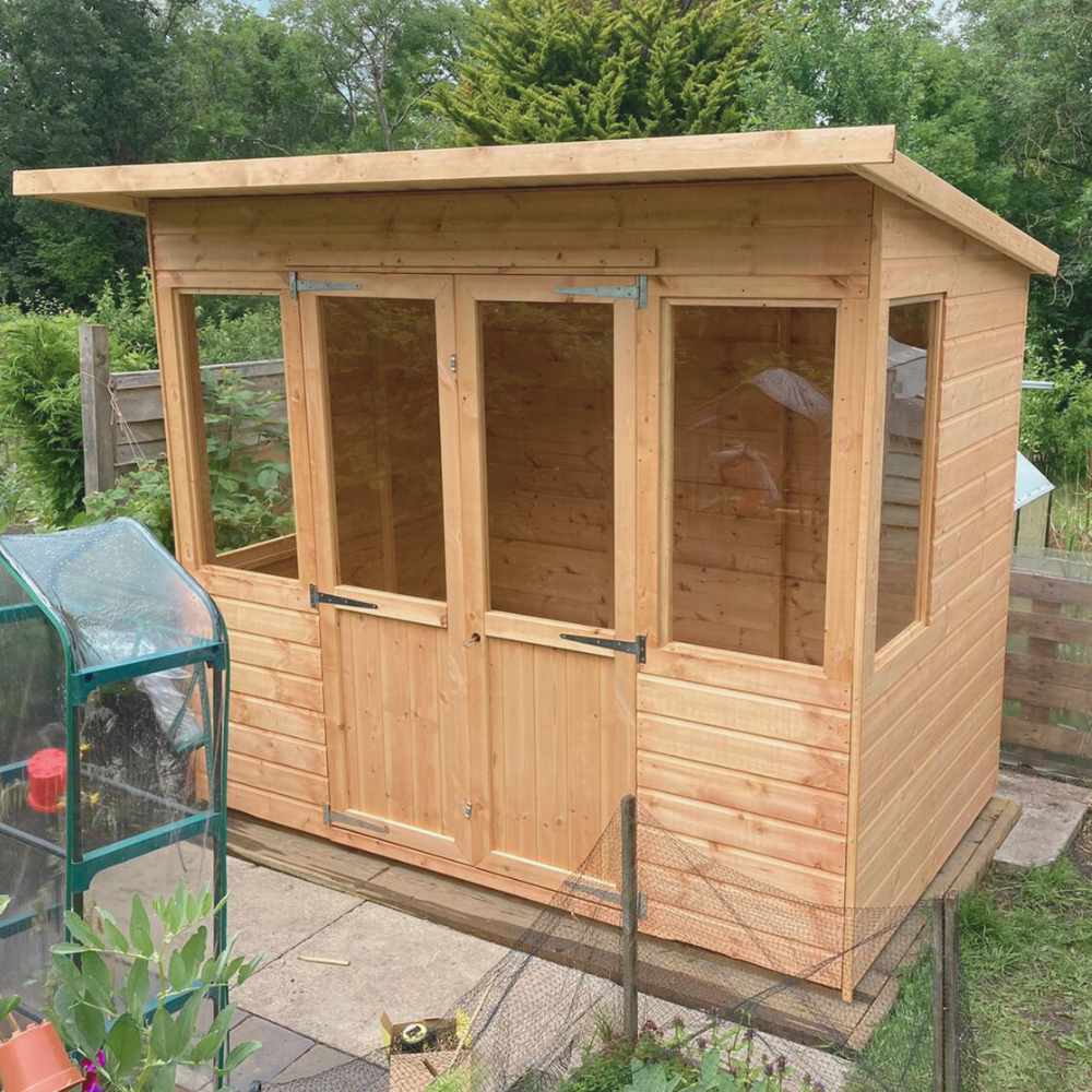 Wooden summerhouse with pent style sloping roof and 4 front windows and two side windows. The two front doors are double opening. The summerhouse is installed on a wooden decking area in a garden.