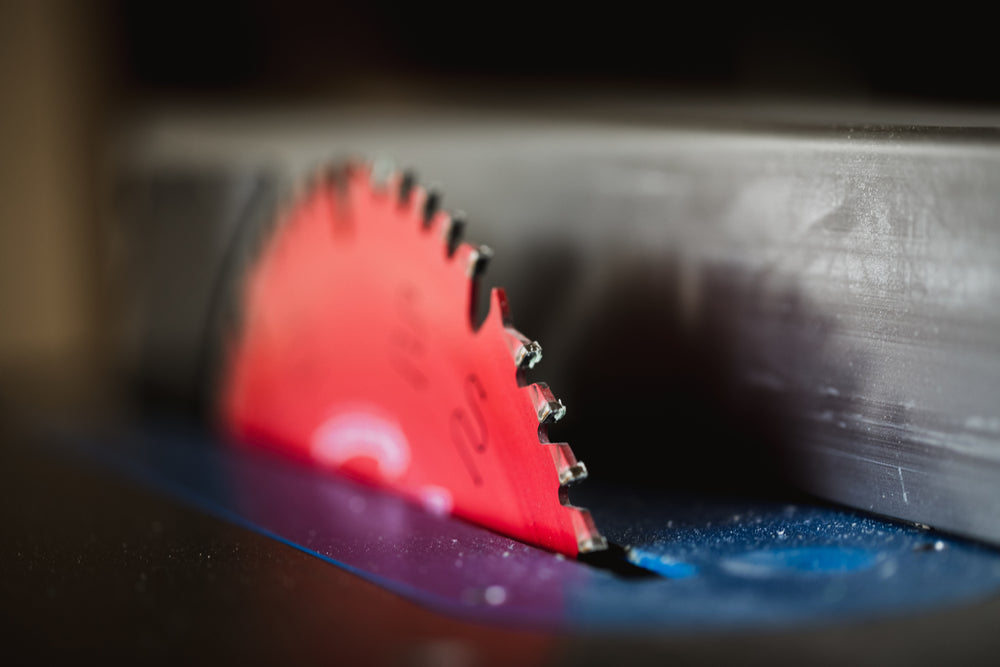 A close-up of a red circular saw blade partially raised above the surface of a table saw, with sharp teeth and a blurred background.