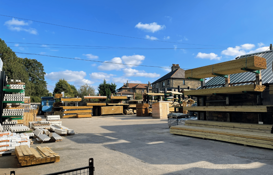 Outdoor lumberyard under a blue sky with scattered clouds. Several stacks of timber and building materials are organized on racks. A few residential houses are visible in the background, surrounded by trees.