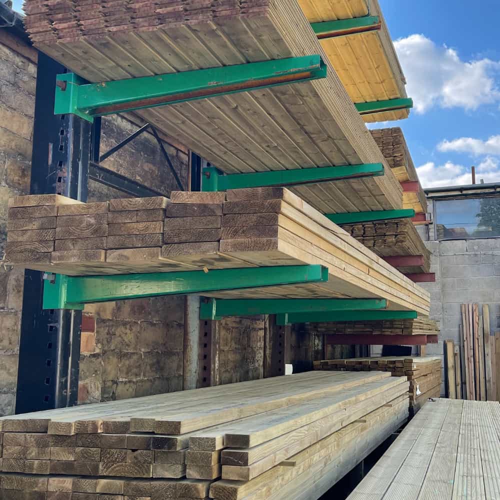Several stacks of wooden planks are neatly arranged on green metal racks outdoors, against a brick wall, with a blue sky and clouds visible in the background.