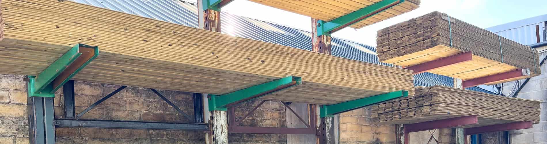 Stacks of wooden planks are neatly arranged on metal racks attached to an outdoor brick wall, each shelf supported by green and red metal beams. A metal roof is visible in the background.